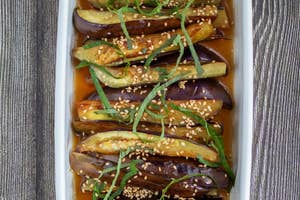 白い皿に盛り付けられたナスの料理。ゴマと刻まれた緑の葉がトッピングされ、ソースがかかっている。