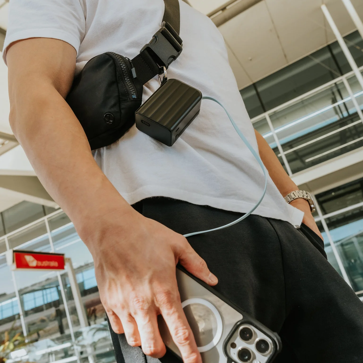Person holding a smartphone with a stand, wearing a white t-shirt and black pants. A small bag is slung across the body. Visible in a shopping setting