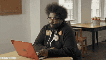 Man in a hoodie sits at a table, tapping rhythmically on a laptop keyboard in a playful manner