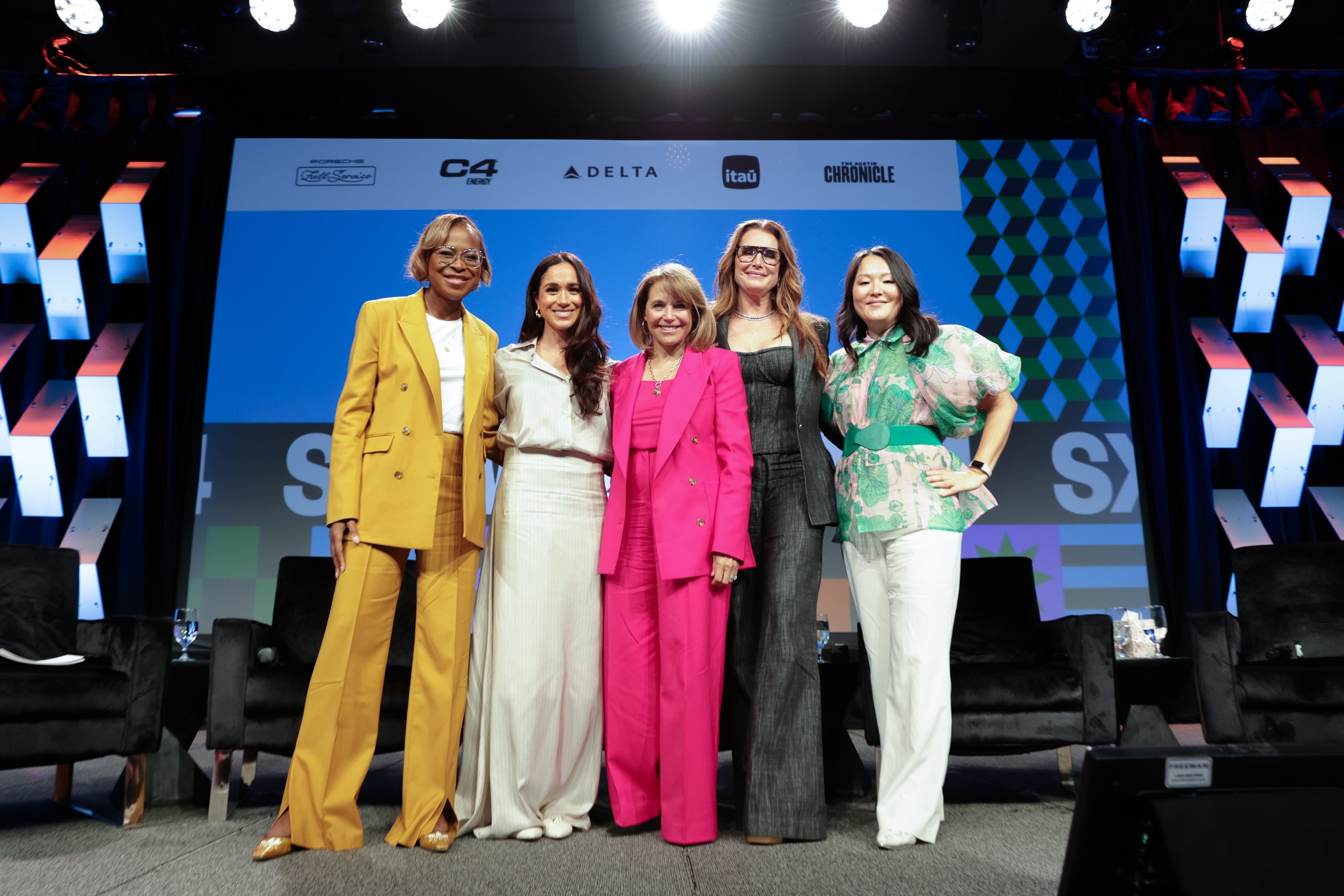 Group of five women smiling on stage at a conference. Each wears distinct, stylish outfits including suits and a floral top