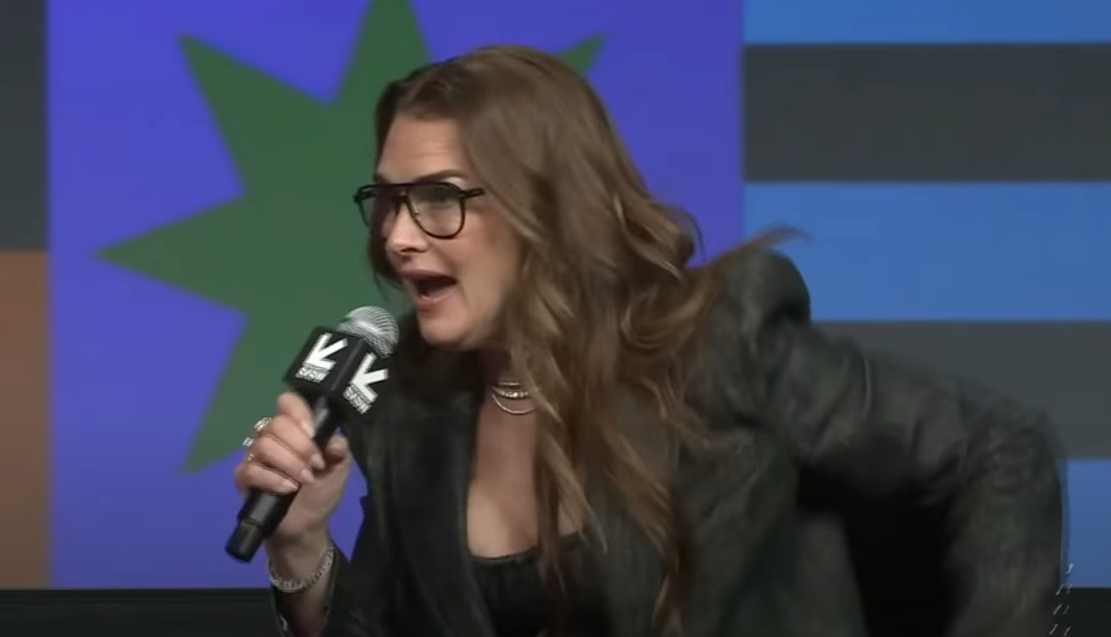 Person speaking passionately into a microphone at an event, wearing a stylish blazer and glasses