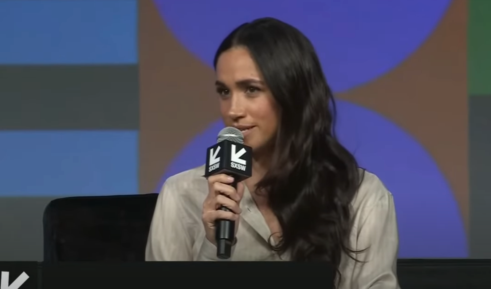 Person speaking into a microphone at a conference, wearing a light-colored blouse, seated on stage