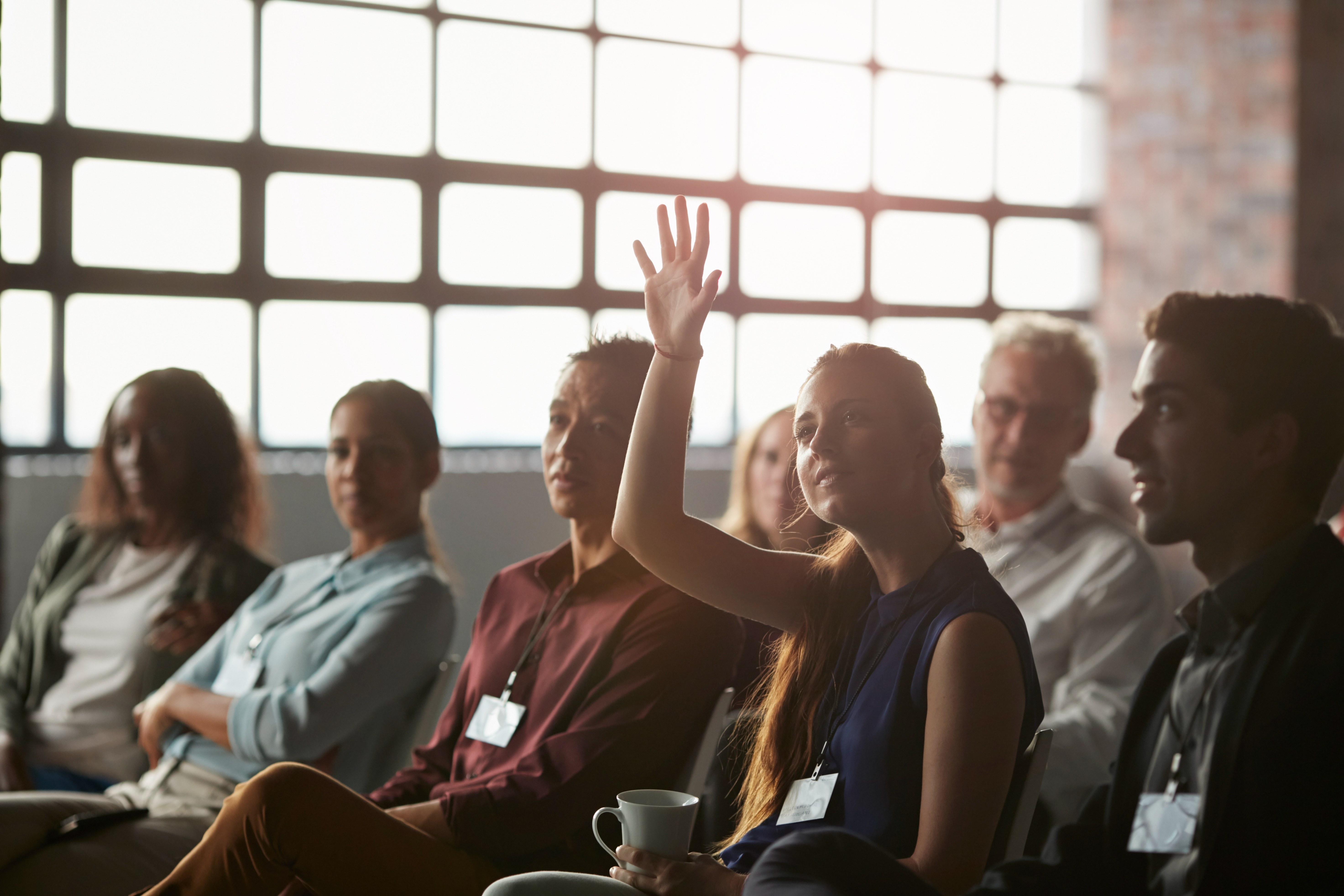 Audience member raising hand during a presentation, holding a mug. Other attendees sit nearby, listening attentively