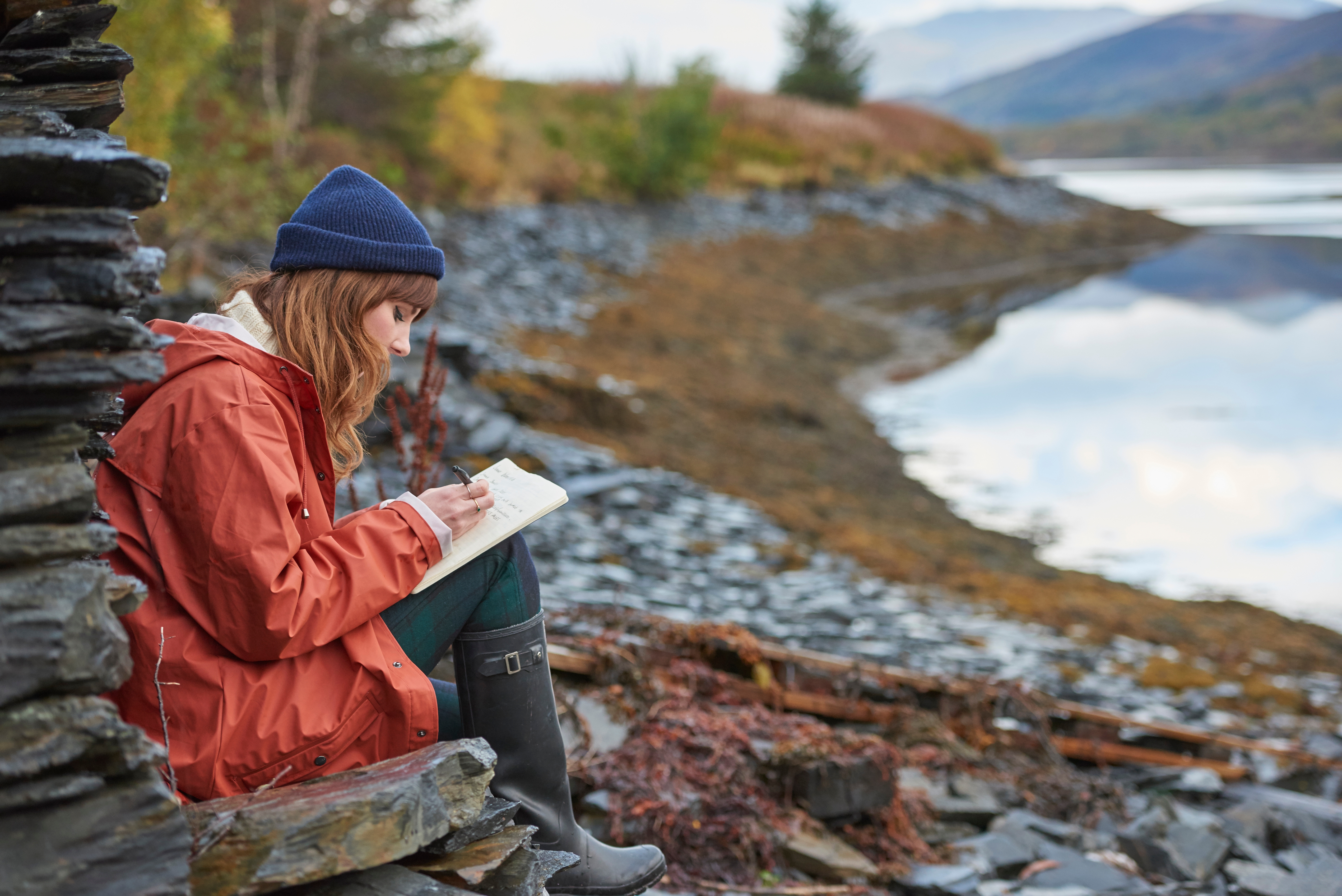 Person in outdoor attire, including a hat and boots, sits on rocks by a lake, writing in a notebook, surrounded by scenic nature