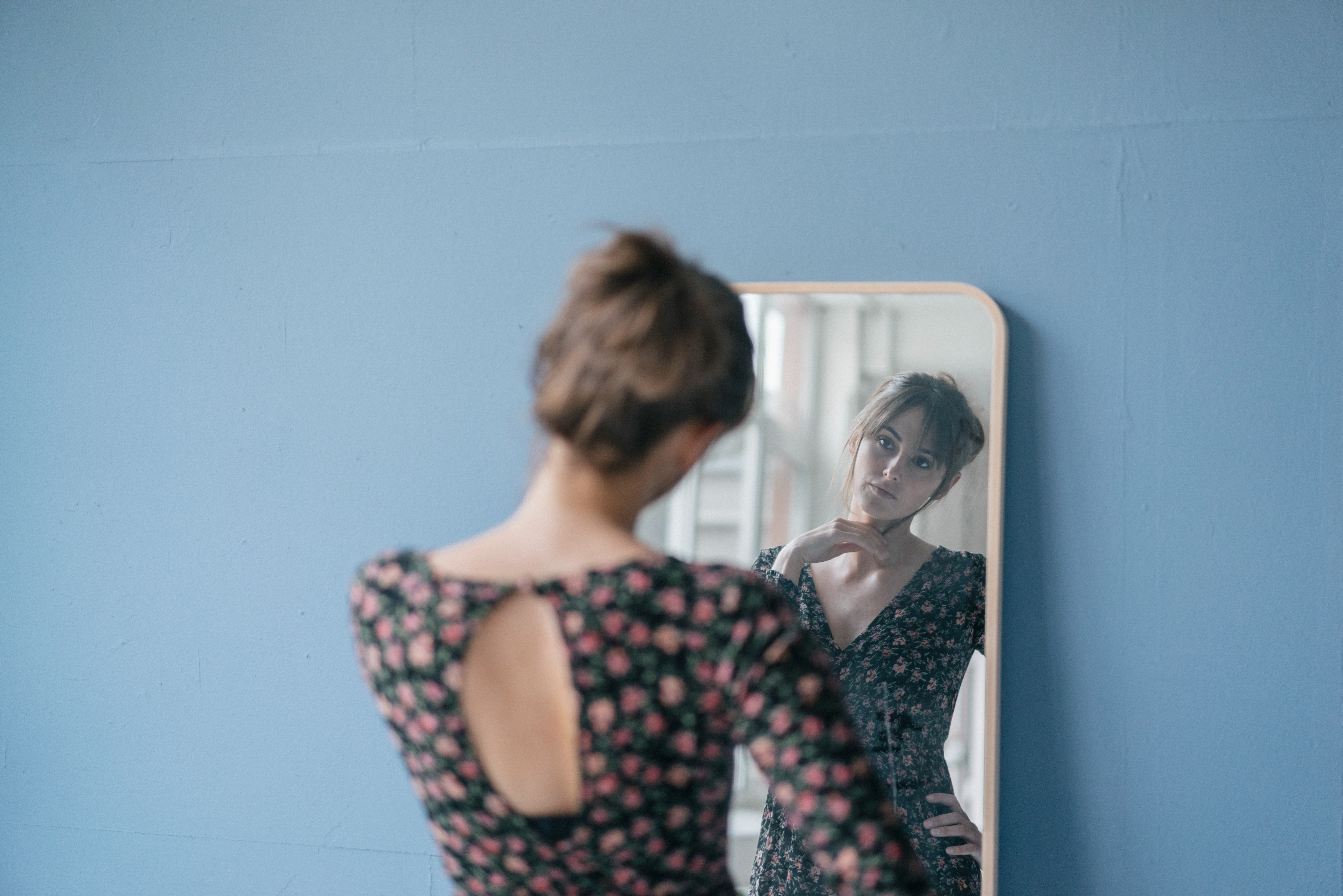 Person in floral dress looks at their reflection in a mirror, standing against a plain wall