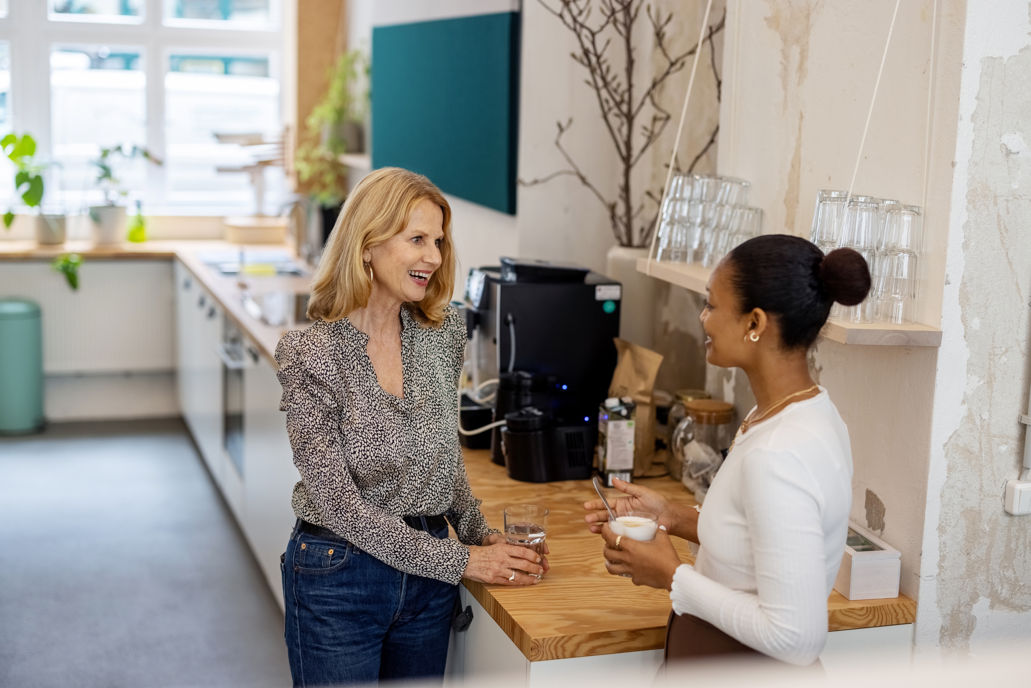 Two women chat in a modern kitchen. One wears a patterned blouse and jeans, the other a white top and apron, both smiling by a coffee machine