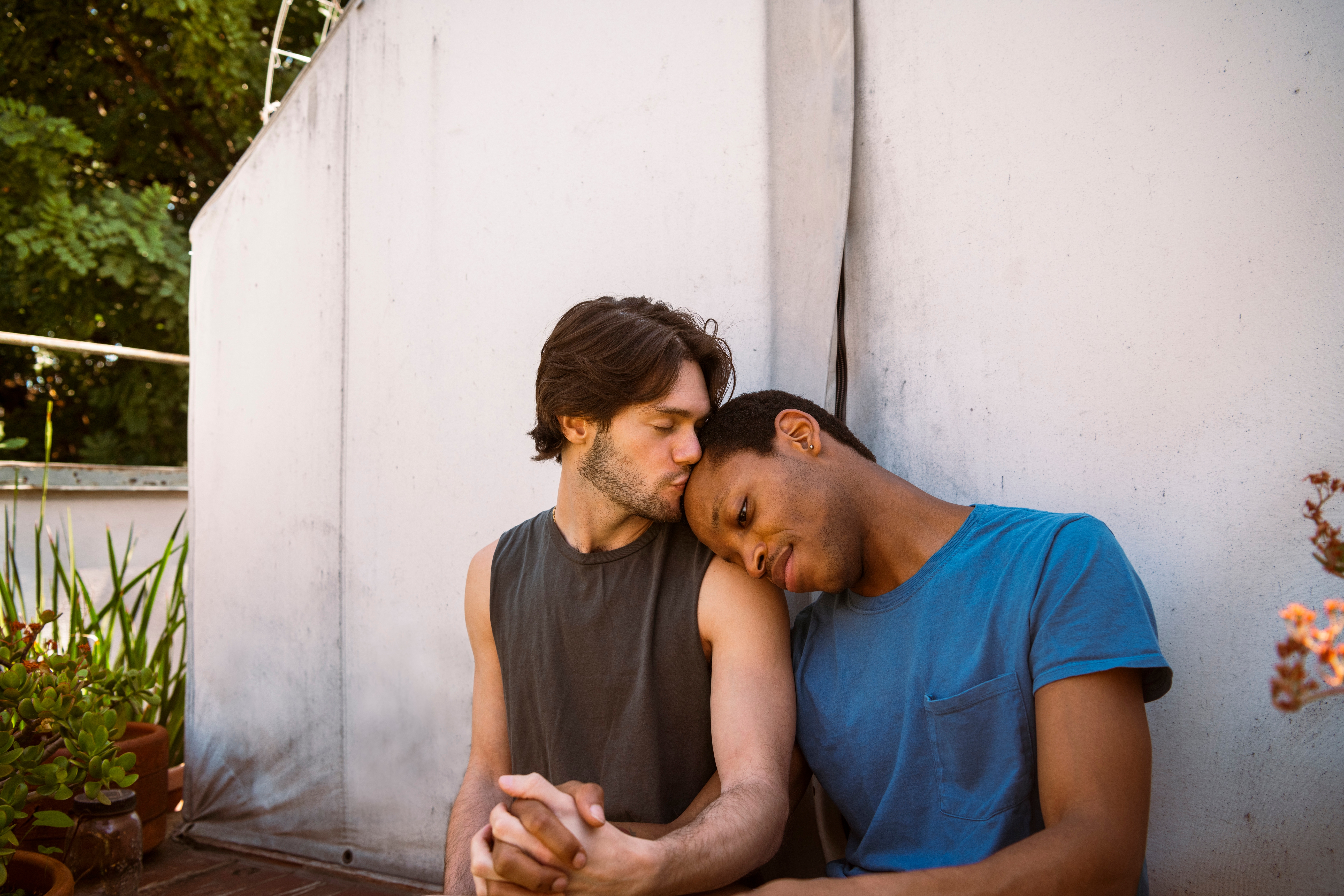 Two people sitting closely, one gently kissing the other's forehead. They appear relaxed and intimate, holding hands on an outdoor patio
