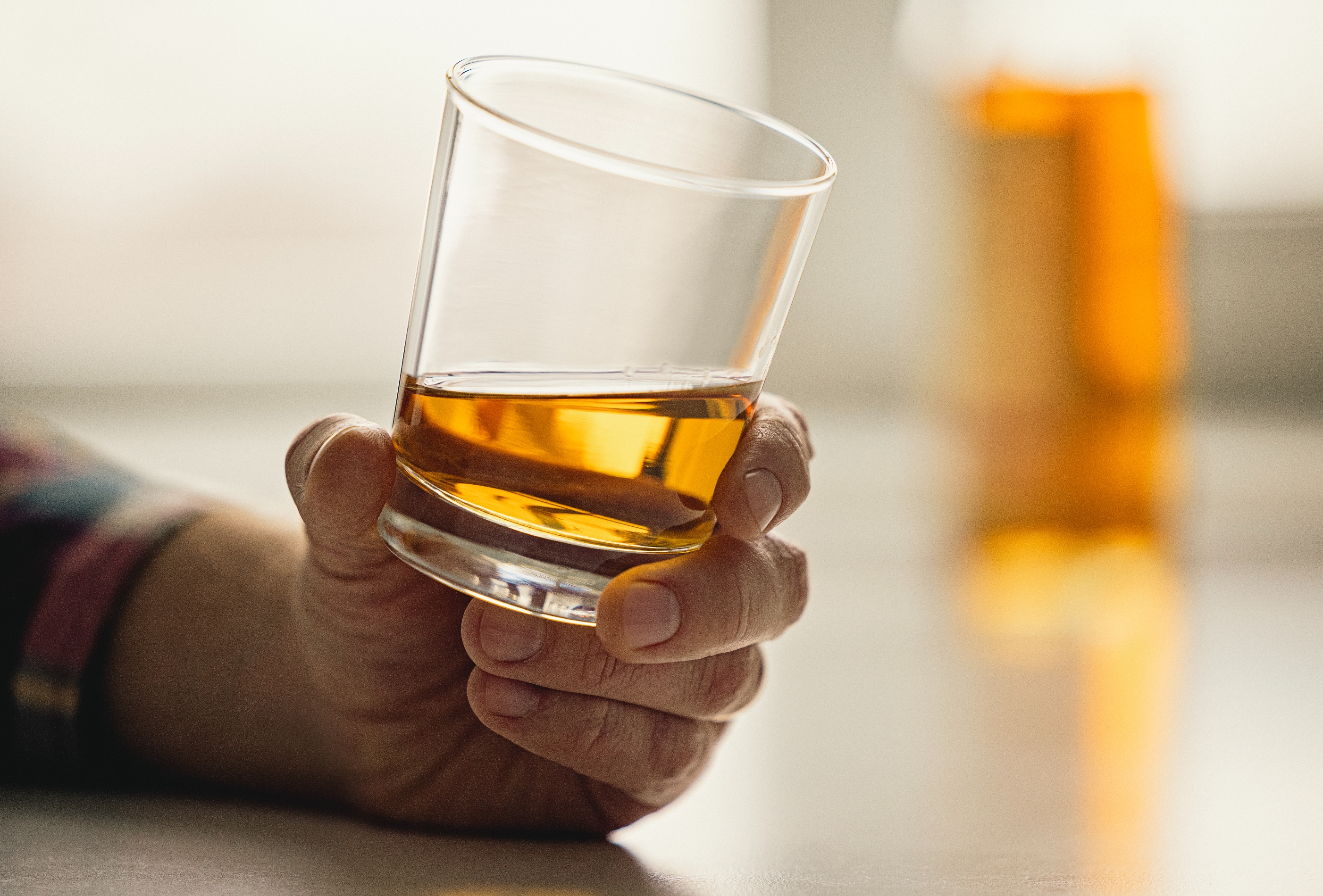 Hand holding a glass of whiskey. Bottle in the background on a table