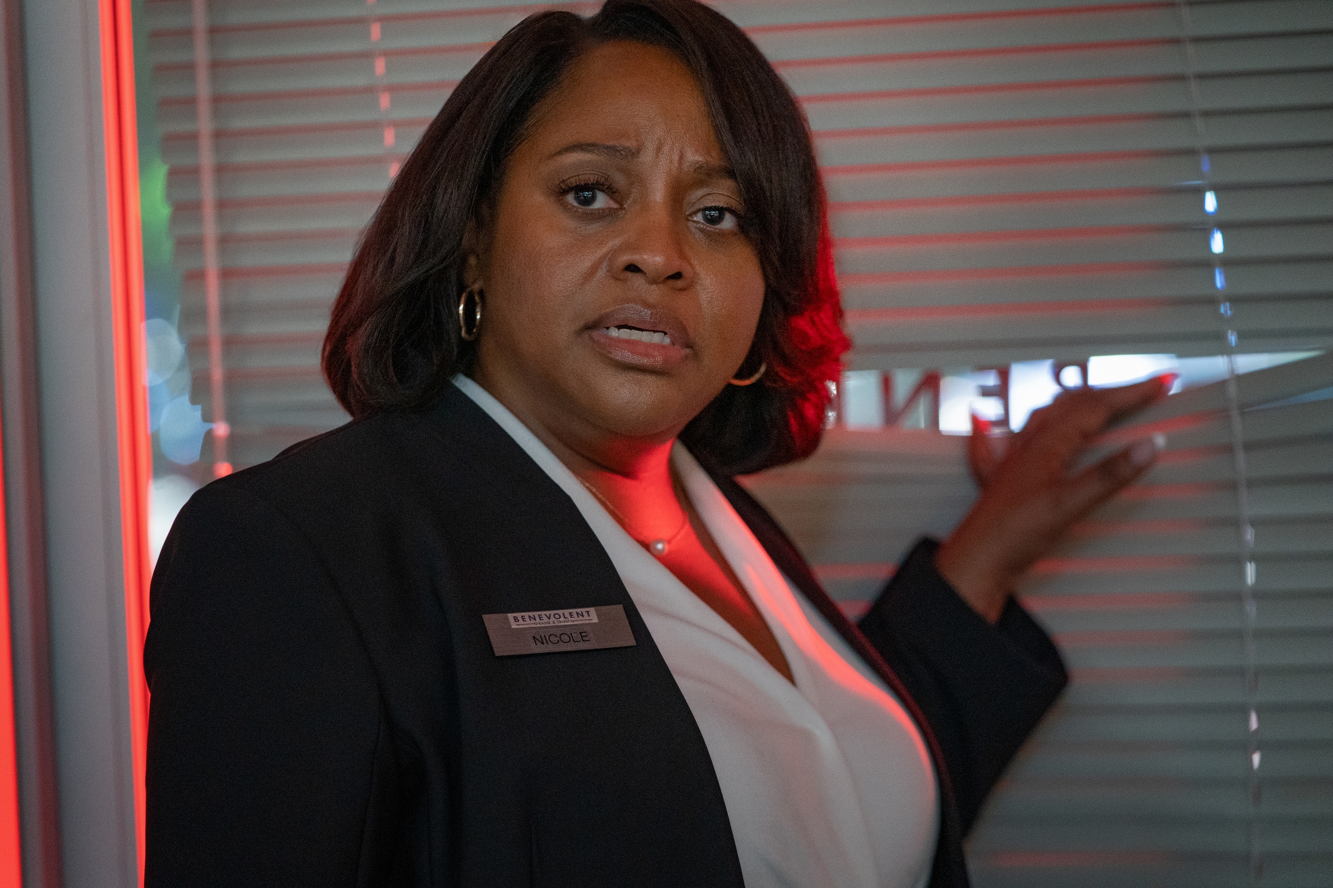 A concerned woman in a blazer stands by window blinds, peering outside, suggestive of a tense scene possibly in a thriller or drama