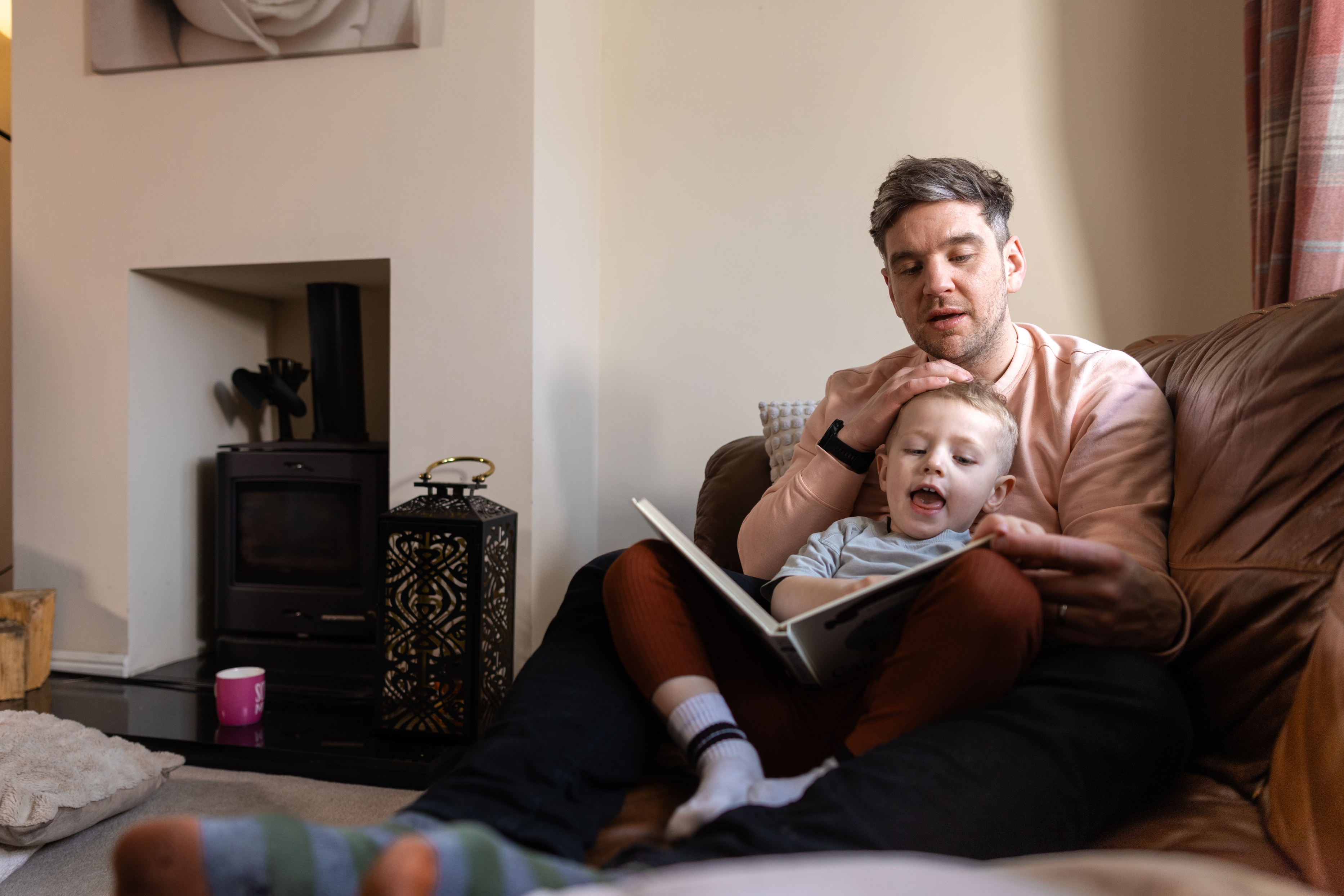 Man reading a book to a young child on a couch in a cozy living room, both looking engaged and relaxed