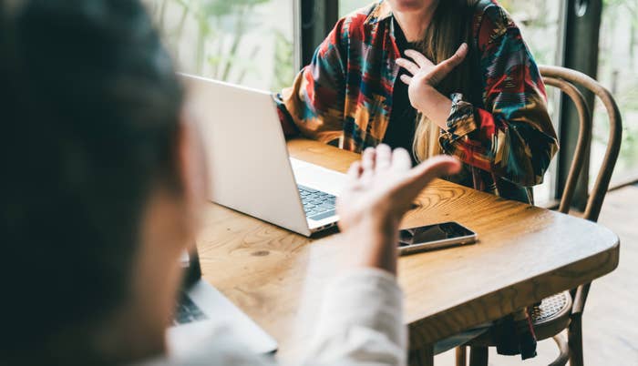 Two people sit at a wooden table with laptops, engaged in a discussion. One gestures while the other listens intently