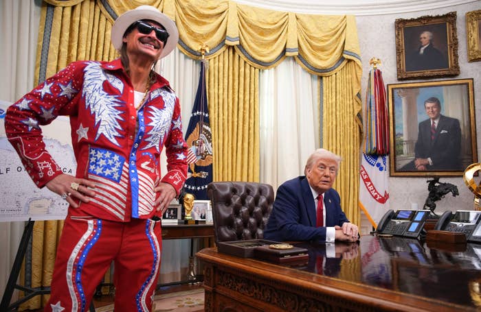 Two men in an ornate office. One in a star-spangled outfit, the other seated in a suit. Portraits and flags in the background