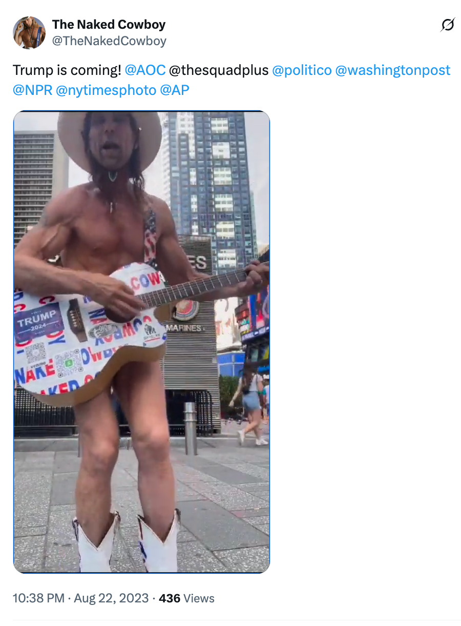 A man wearing a cowboy hat, boots, and briefs plays a guitar covered in political stickers in a busy city setting