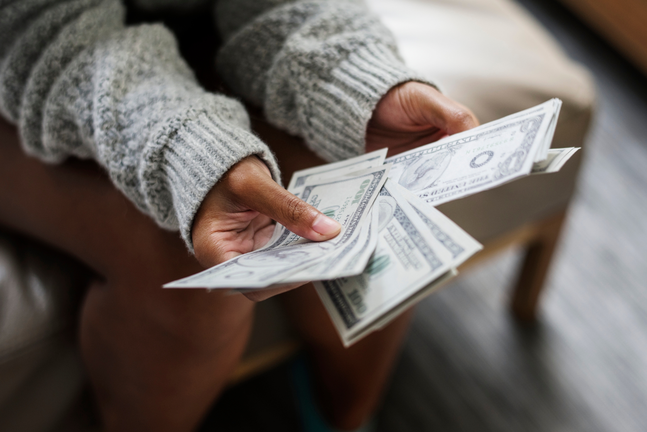 Person sitting, counting multiple 100-dollar bills while wearing a cozy sweater