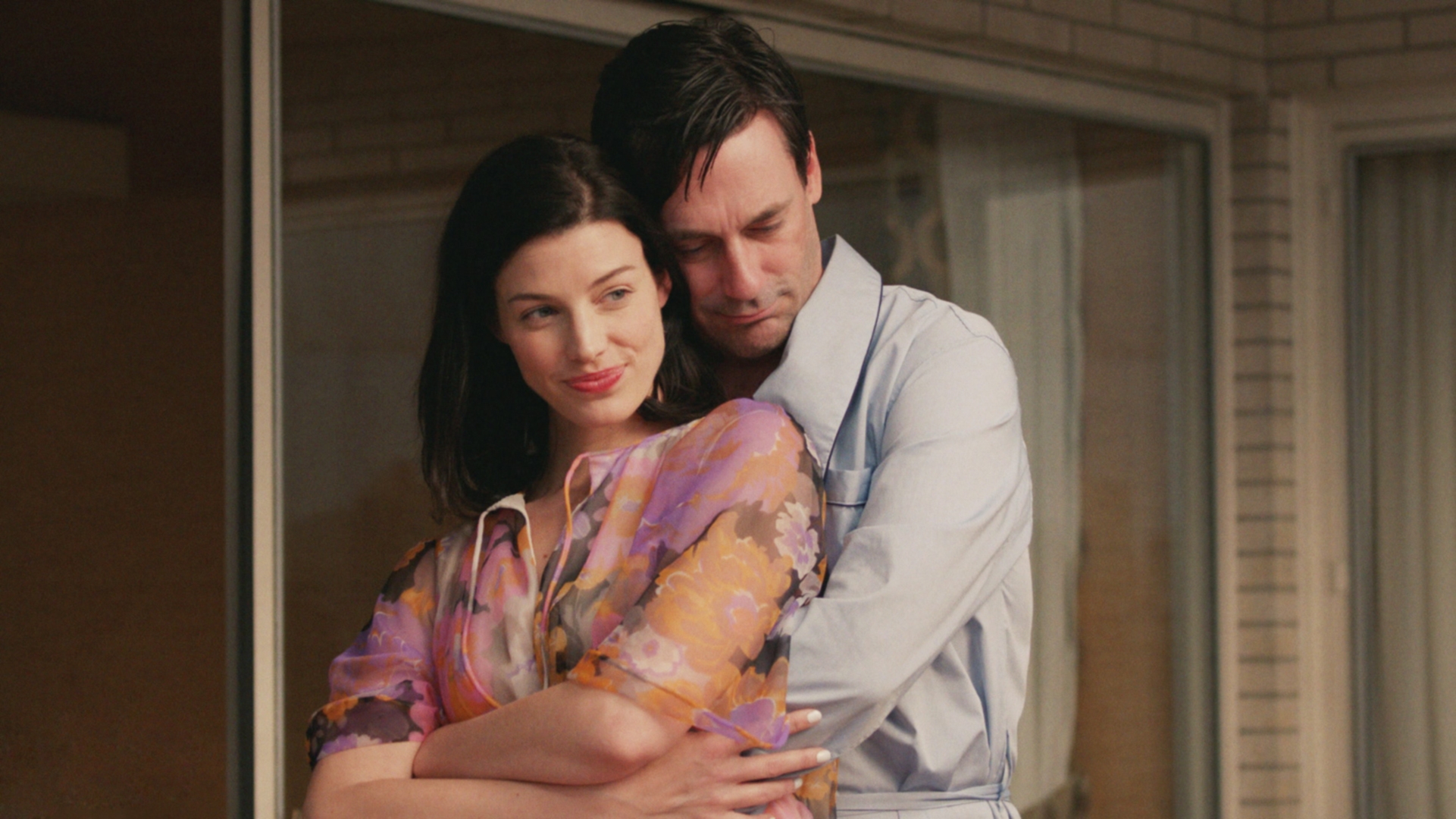 A couple embraces affectionately on a porch. The woman wears a floral dress, while the man dons a light-colored shirt