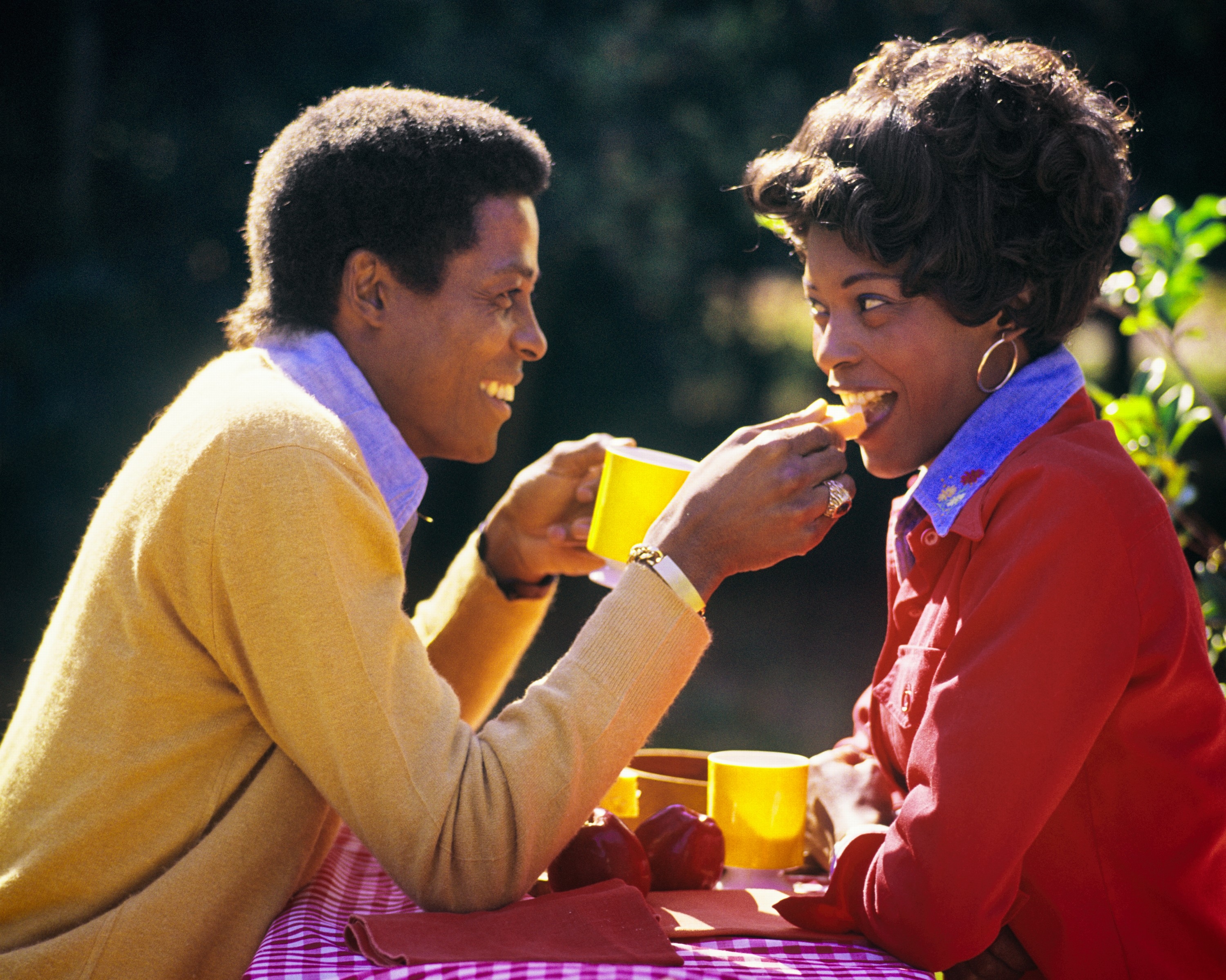 A couple laughs and shares a meal outdoors, seated at a picnic table. They gaze lovingly into each other's eyes, holding bright cups