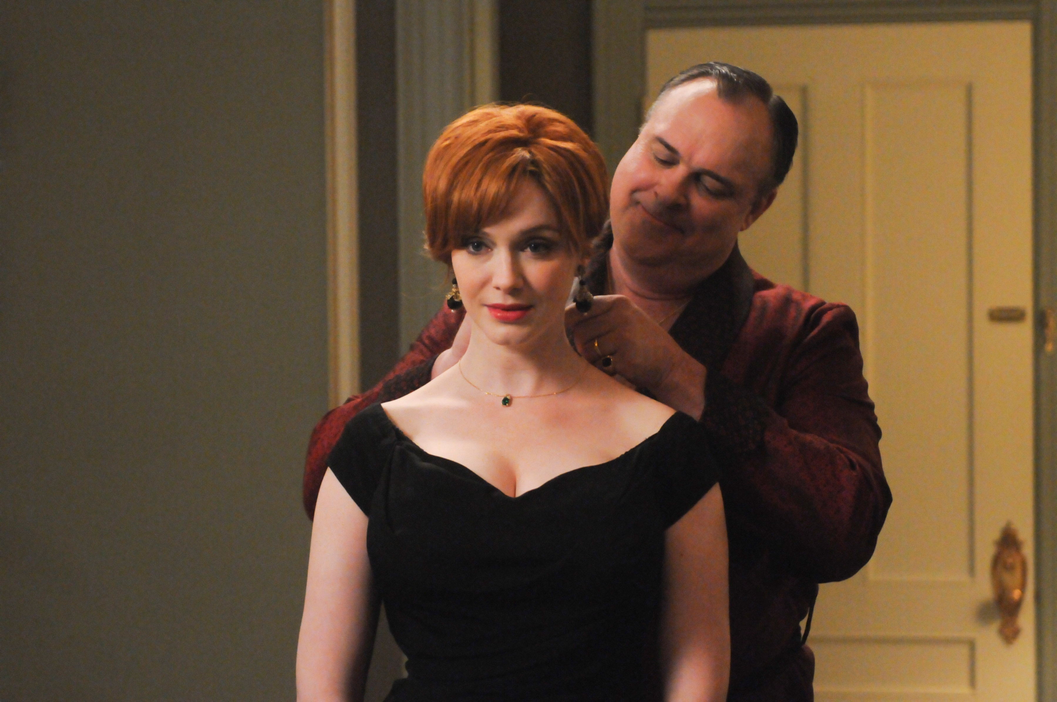 A person helps another person adjust a necklace. The front person wears a vintage-inspired black dress. The setting is indoors