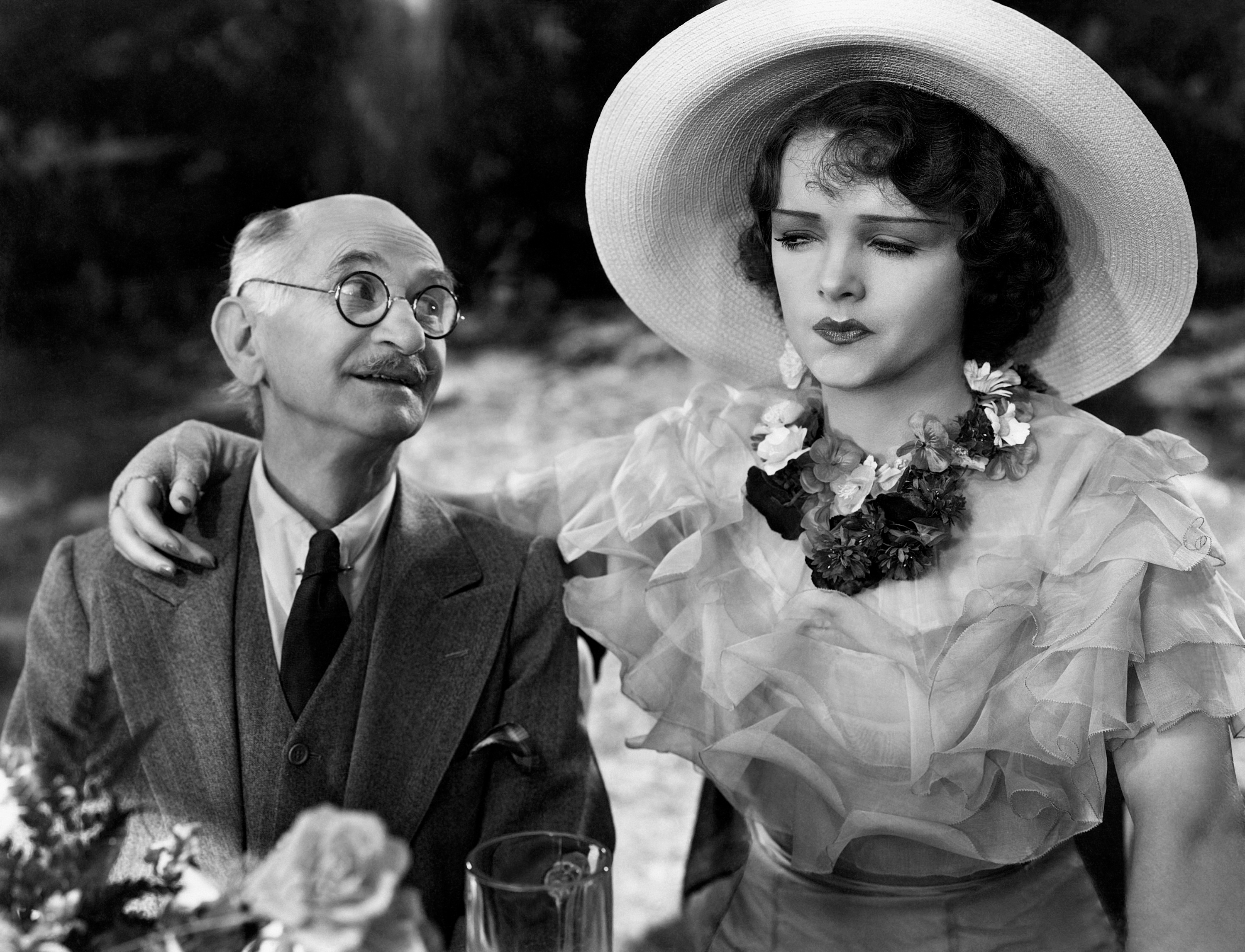 A vintage couple, man in a suit and glasses, woman in a ruffled dress and large hat, sit closely with a thoughtful expression, evoking romance