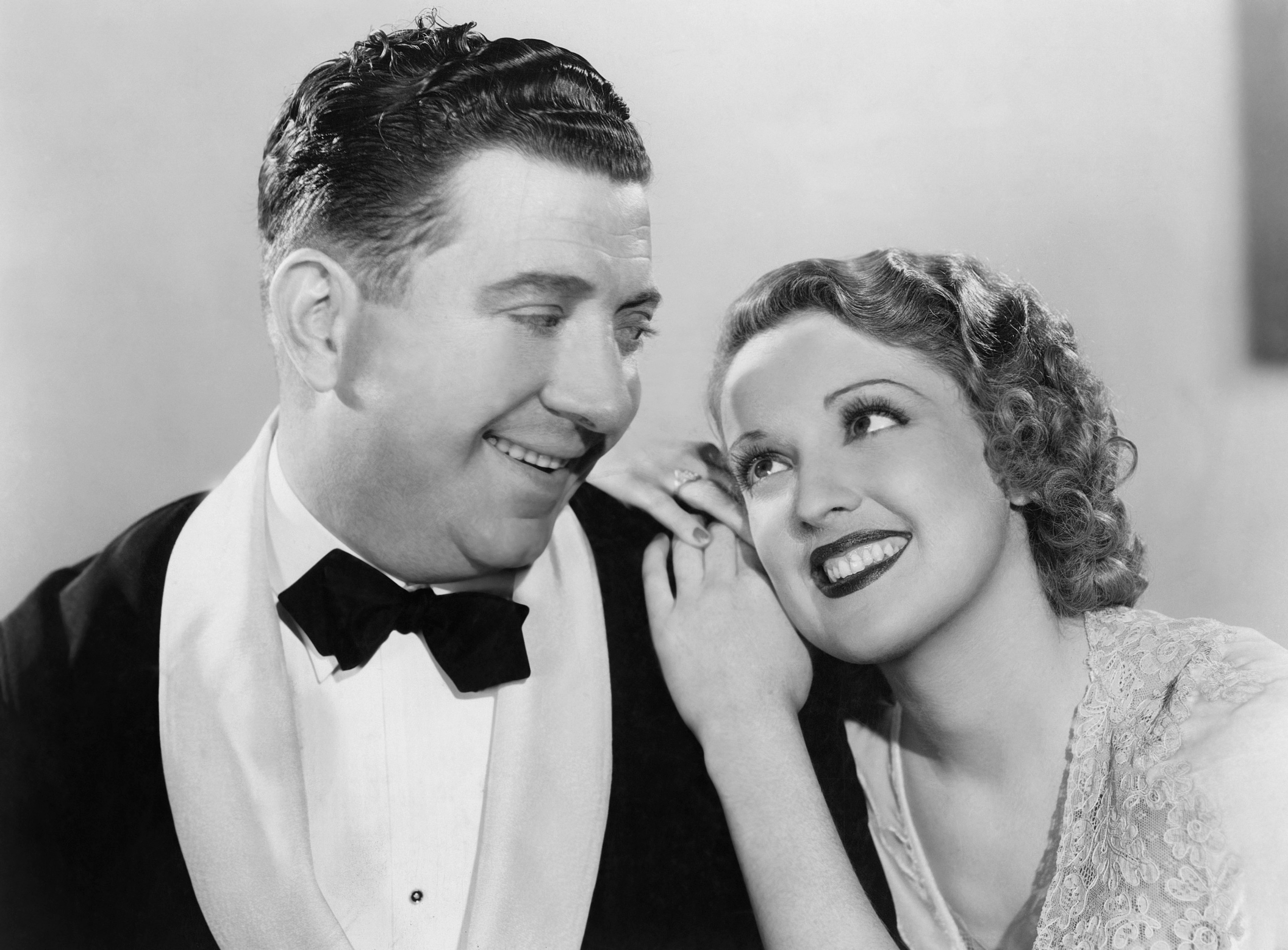 A couple smiles warmly at each other. The man wears a tuxedo, and the woman leans on his shoulder with a lace dress, exuding affection and joy