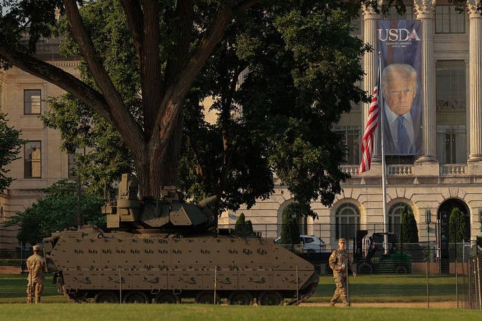 Military personnel and armored vehicle in front of government building with large banner of a public figure