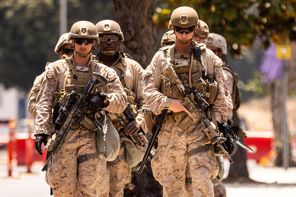 Soldiers in tactical gear and helmets walk in formation, each carrying rifles. They appear focused and alert, suggesting a military operation or exercise