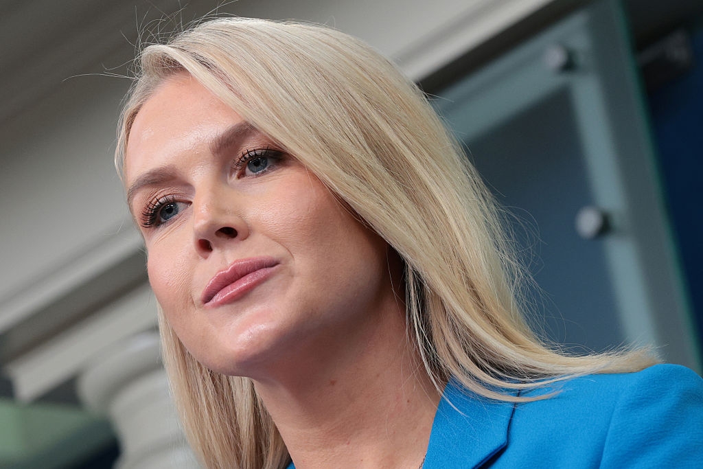 A woman with long blonde hair in a blue blazer stands confidently, likely at a press event