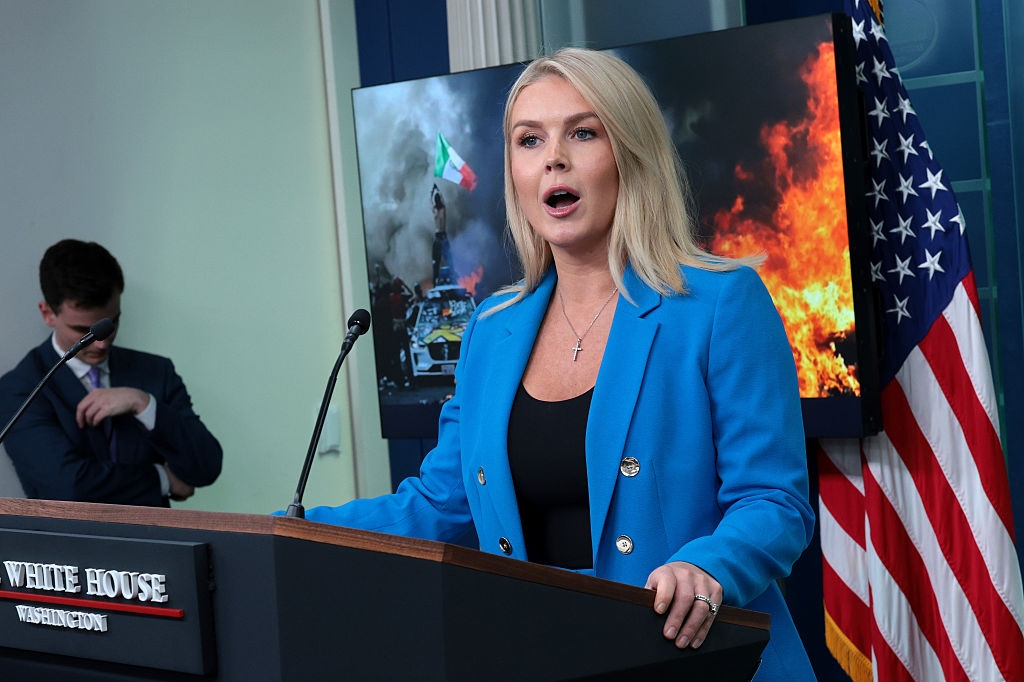 A woman in a blue blazer speaks at a podium with a fire and flag on screen behind her, in front of the U.S. flag