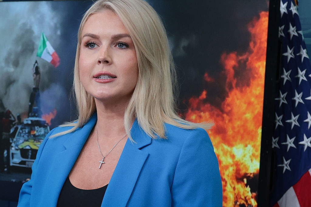 A woman in a blue blazer speaks in front of images depicting fire and an Italian flag, alongside a U.S. flag
