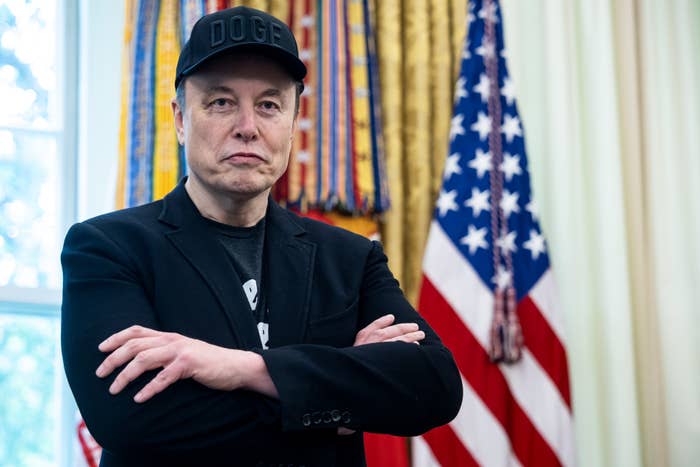 Elon Musk standing withhis  arms crossed, wearing a casual cap and blazer, in a formal room with a U.S. flag in the background