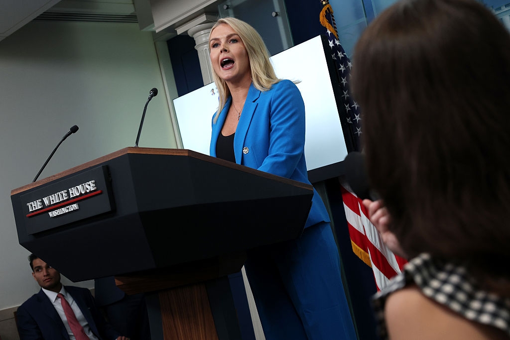 A woman in a blue suit speaks at a podium labeled "The White House" during a press briefing, while reporters listen and take notes