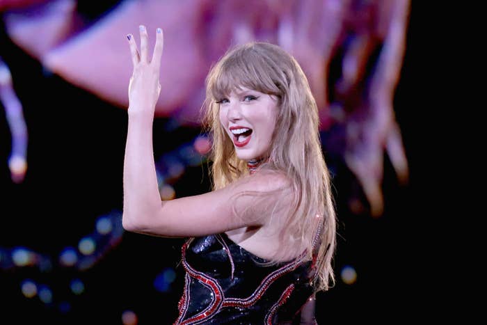 Taylor Swift on stage wearing a shimmering outfit waves and smiles energetically during a performance