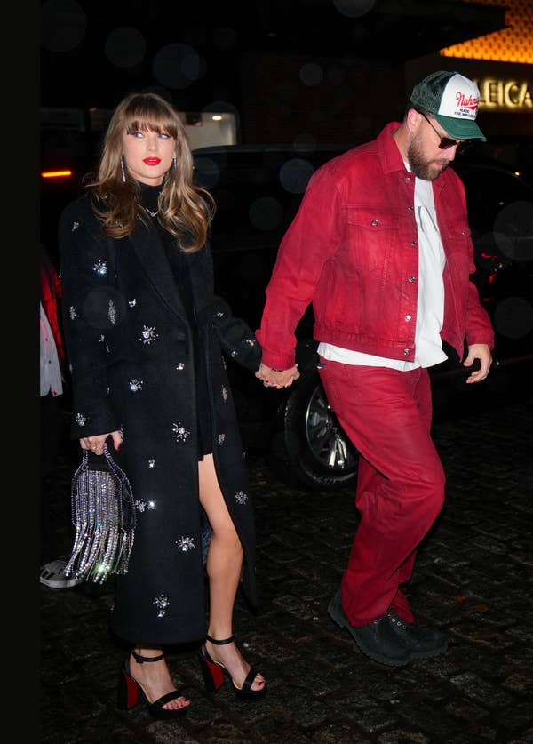 Taylor Swift in a long sparkly coat and matching fringe bag holding hands with Travis Kelce