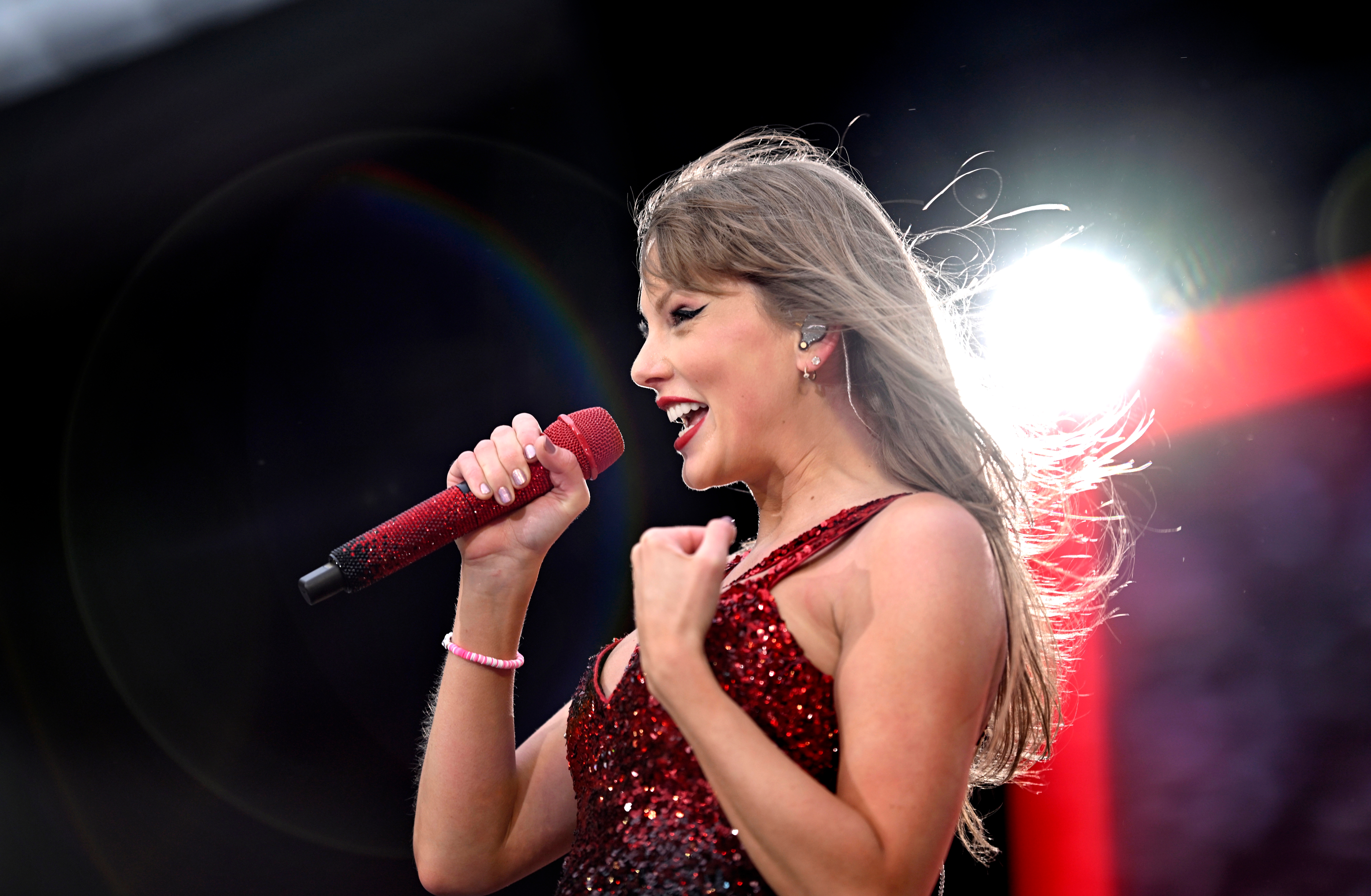 Taylor Swift singing energetically on stage in a sparkling sequined outfit, holding a red microphone with a backdrop of stage lights
