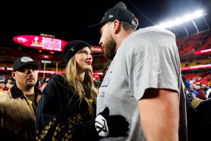 Taylor Swift in a hat and hooded sweatshirt talking closely with Travis Kelce at a football game