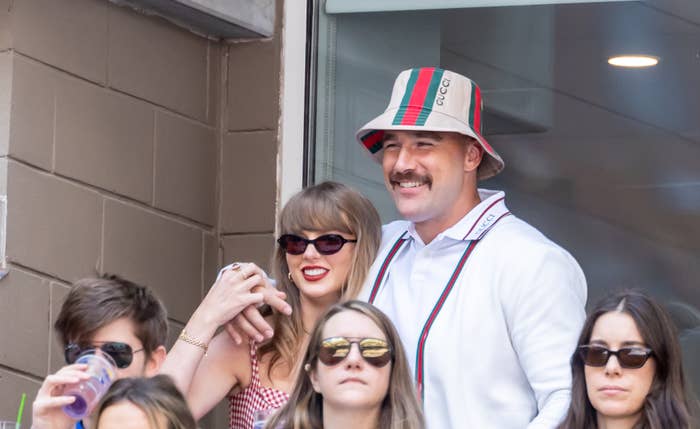 Travis Kelce wears a striped bucket hat with an arm around Taylor Swift at a sporting event