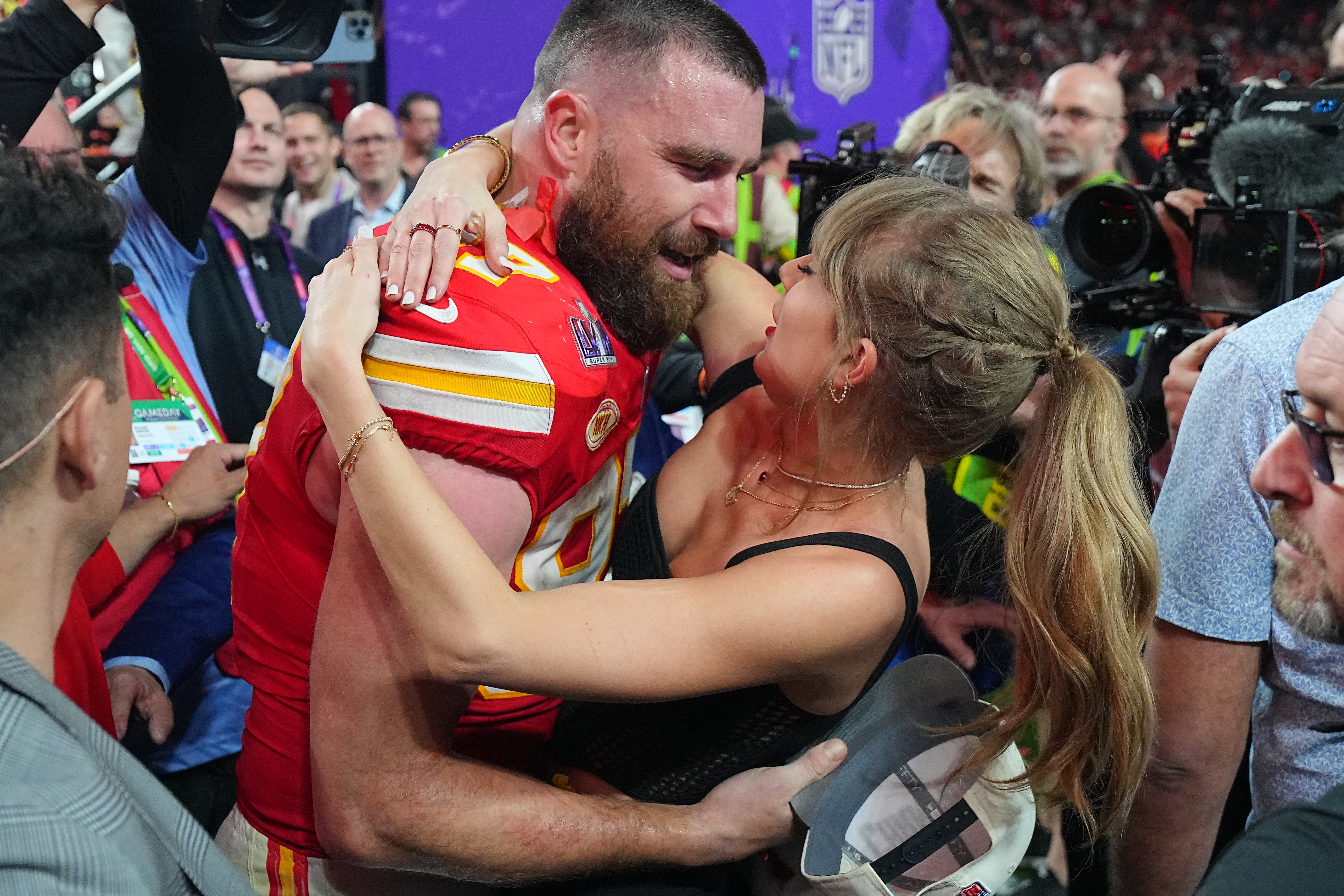 Travis Kelce in gear celebrates with Taylor Swift in a chic dress on a crowded sports field, surrounded by fans and media
