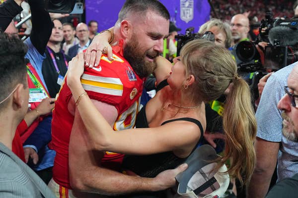 Travis Kelce in gear celebrates with Taylor Swift in a chic dress on a crowded sports field, surrounded by fans and media