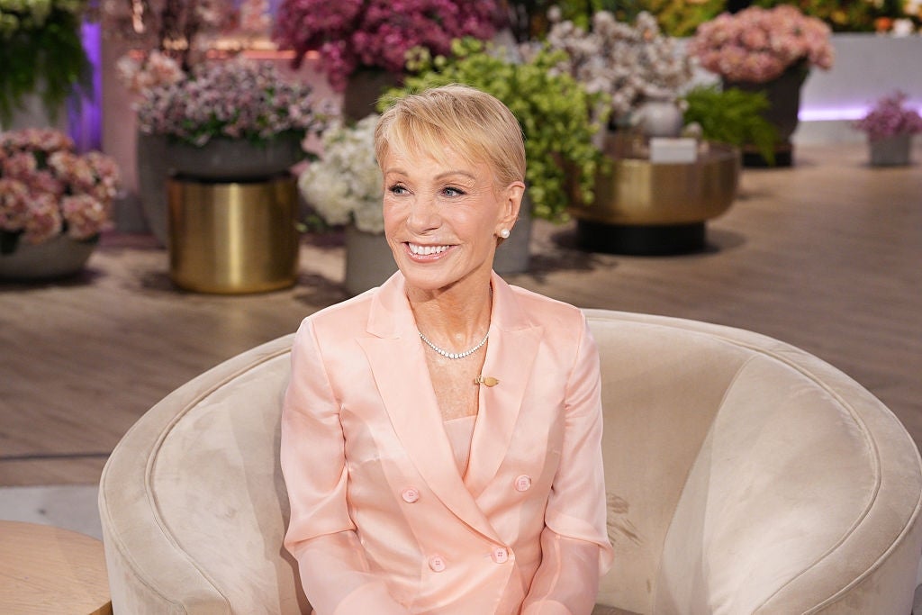 A person sits on a cream chair in a bright room decorated with flowers, wearing a light-colored suit and a necklace, smiling