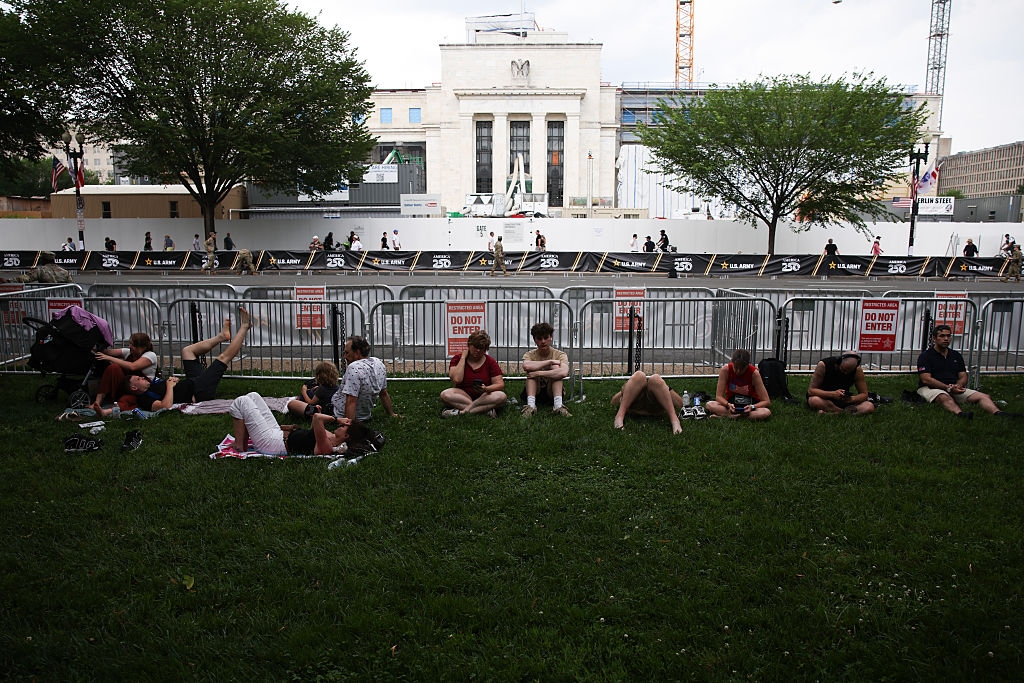 People are sitting and lying on a grassy area outside, near a fenced-off construction site with a prominent building in the background