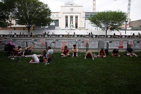 People are sitting and lying on a grassy area outside, near a fenced-off construction site with a prominent building in the background