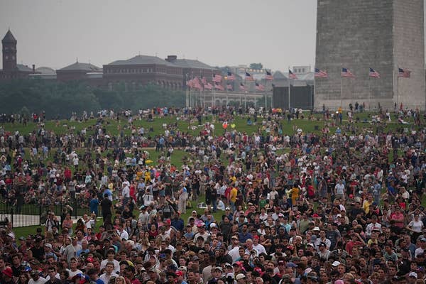 Large crowd gathered on a green lawn near the Washington Monument, with visible flags and city buildings in the background