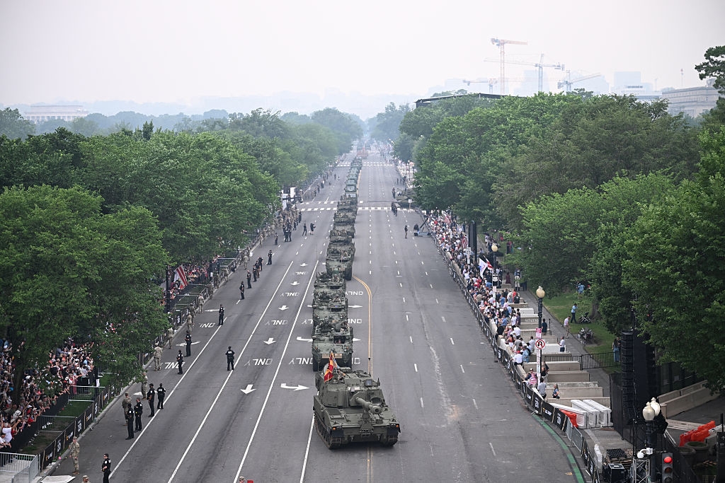 Military tanks line a city street during a parade, with spectators watching from the sidewalks