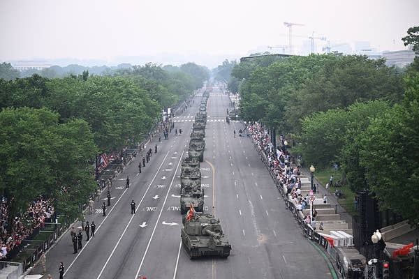 Military tanks line a city street during a parade, with spectators watching from the sidewalks