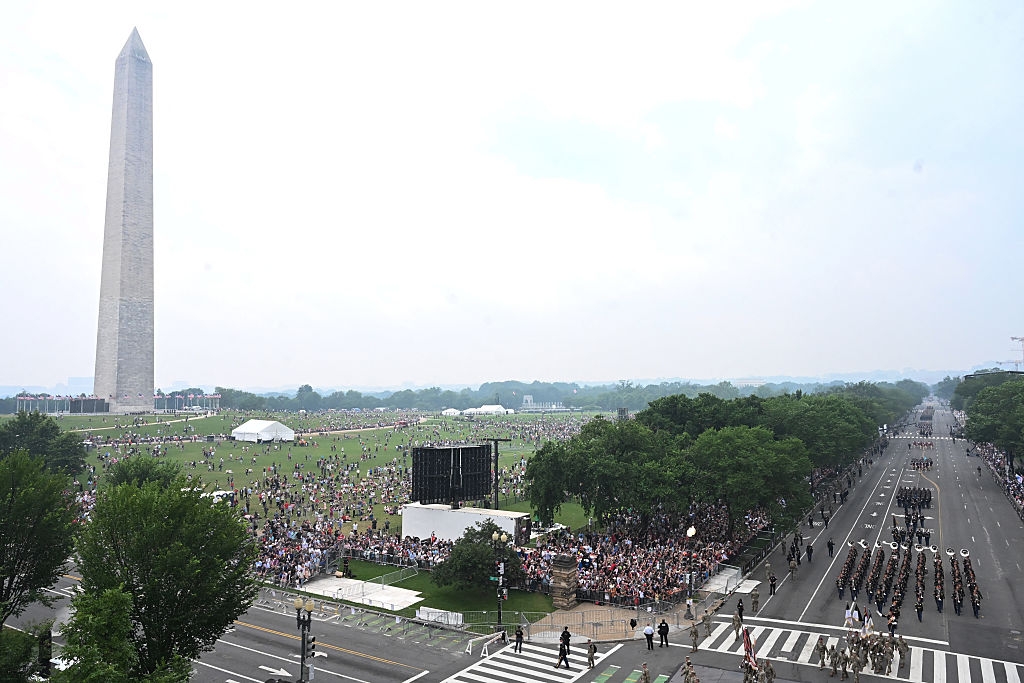 Large crowd gathered near the Washington Monument, with a parade and military personnel marching on a road, trees lining the area