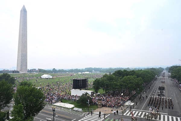 Large crowd gathered near the Washington Monument, with a parade and military personnel marching on a road, trees lining the area