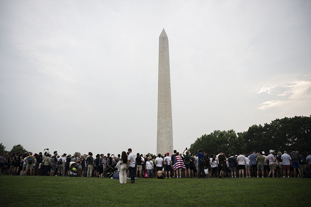 Crowd gathers at the base of the Washington Monument on a cloudy day, some capturing photos