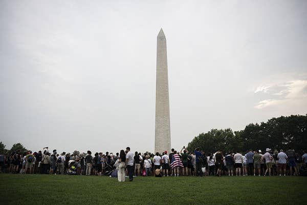 Crowd gathers at the base of the Washington Monument on a cloudy day, some capturing photos