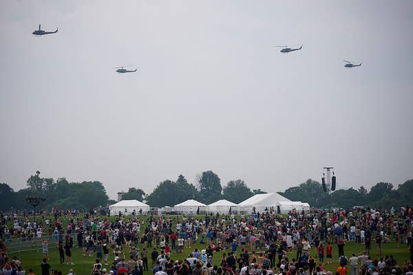 Helicopters fly over a large outdoor gathering with crowds and tents below, likely a public event or festival
