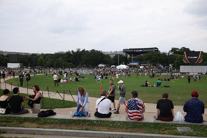 People sitting and walking in a large grassy park area, gathered for an outdoor event with tents and screens