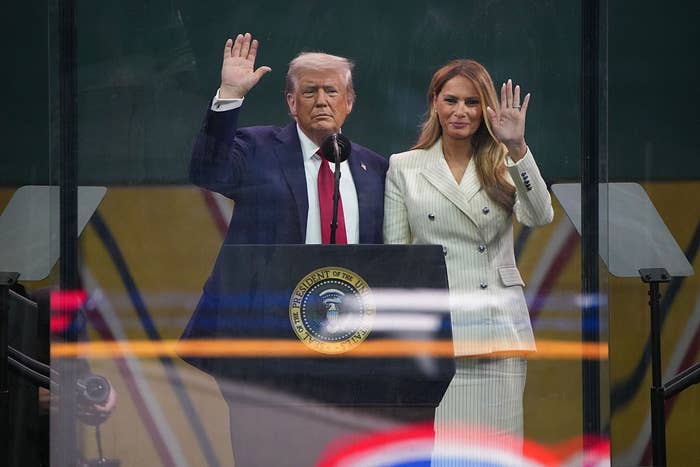 Two people stand at a podium, waving. The man wears a suit and red tie; the woman wears a stylish pinstriped suit